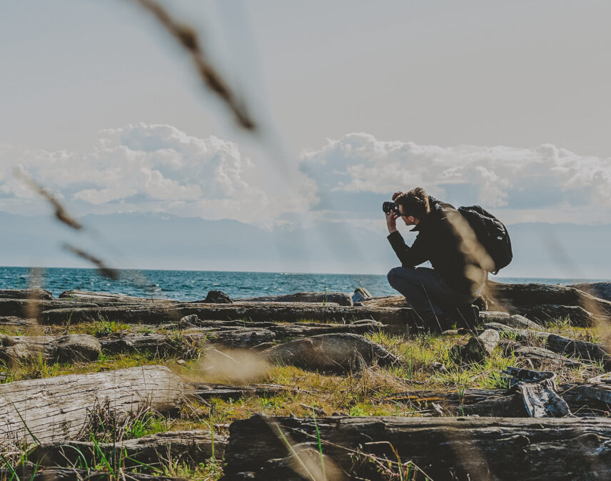 Photographer capturing coastal scenery in Colwood, near Victoria, BC, with driftwood, grassy terrain, and ocean waves under a cloudy sky