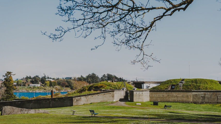 Historic fort surrounded by green grass and trees in Colwood, near Victoria, BC, overlooking the ocean with distant buildings and benches in a serene park setting