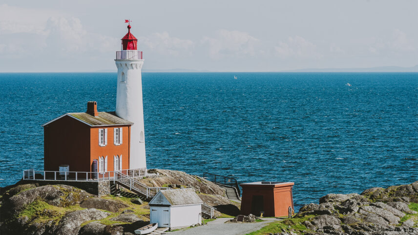 Historic red and white lighthouse overlooking the ocean in Colwood, near Victoria, BC, set on rocky terrain with a clear blue sky and distant sailboats in the background