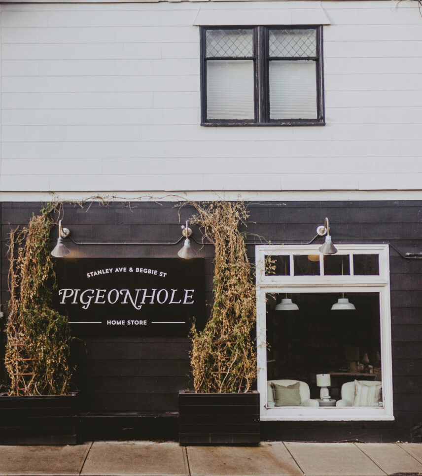 Exterior of Pigeonhole Home Store at Stanley Ave & Begbie St, featuring black siding, large window display with cozy furniture, and decorative plants