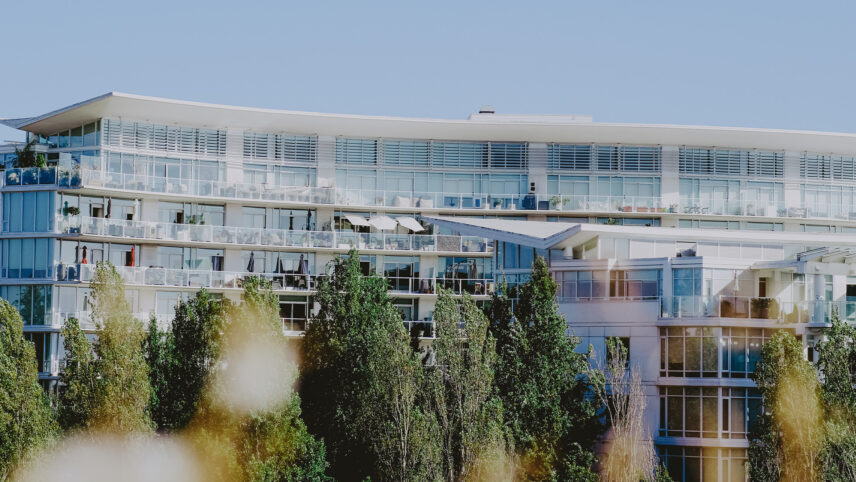 Modern multi-story condominium building with large glass windows and private balconies, surrounded by lush green trees