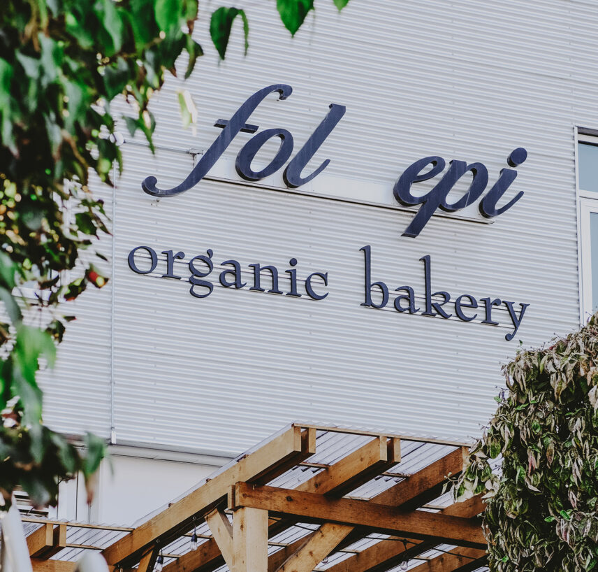 Exterior signage of Fol Epi organic bakery on a modern metal-clad building with wooden pergola and greenery