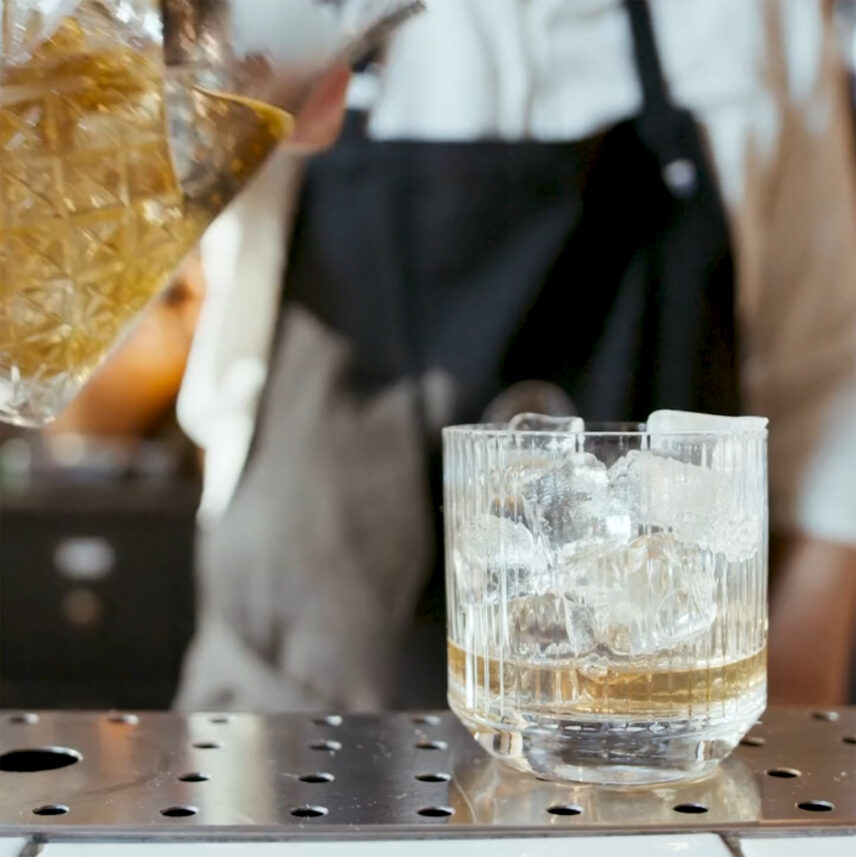 Bartender pouring a golden beverage into a glass filled with ice, placed on a metal counter