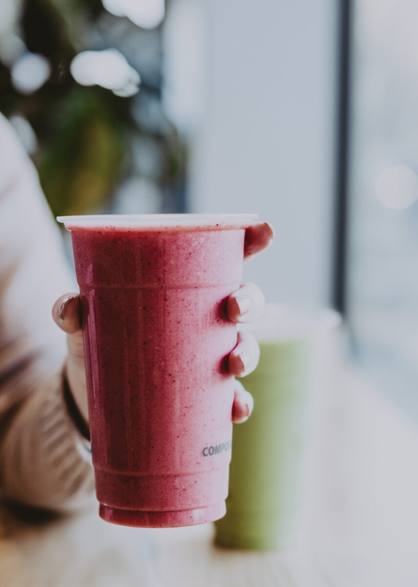 Hand holding a red smoothie in a compostable cup with a green smoothie in the background on a wooden table