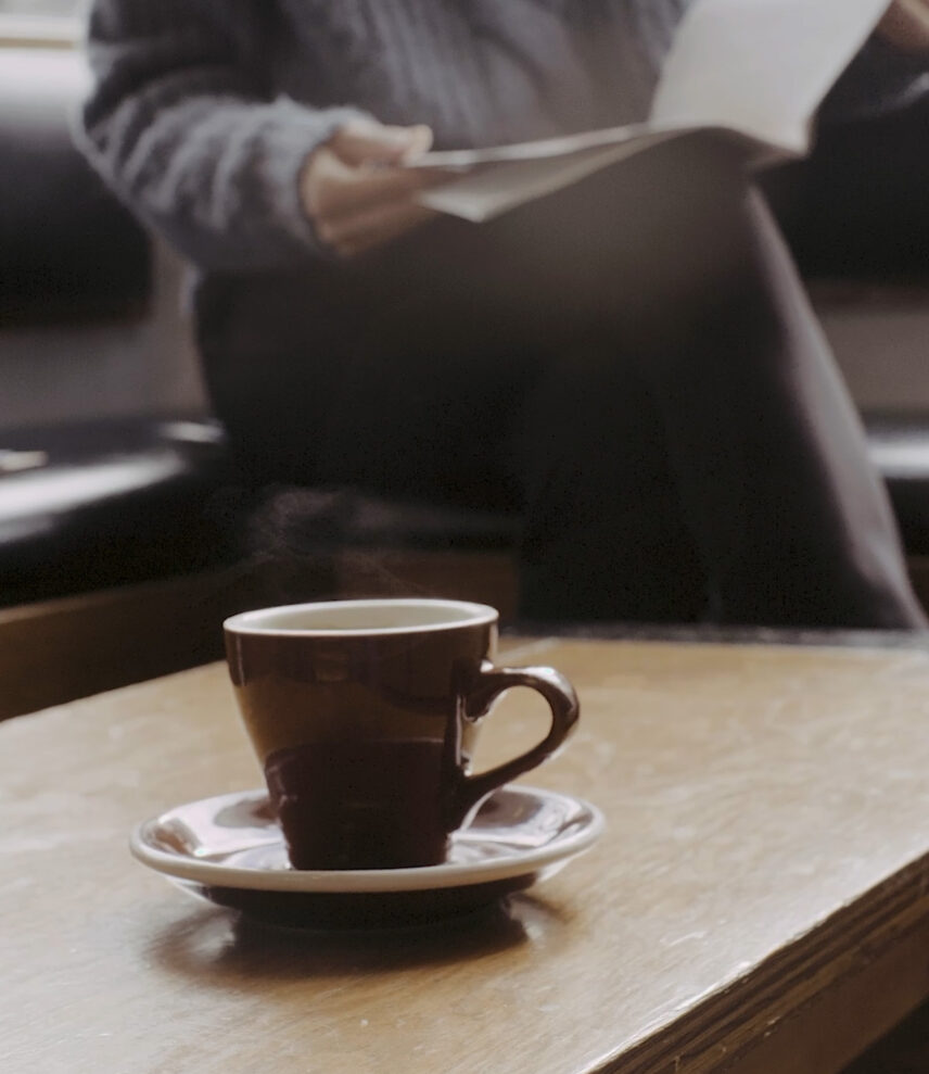 A steaming cup of coffee in a brown ceramic mug on a saucer, placed on a wooden table, with a person reading in the background
