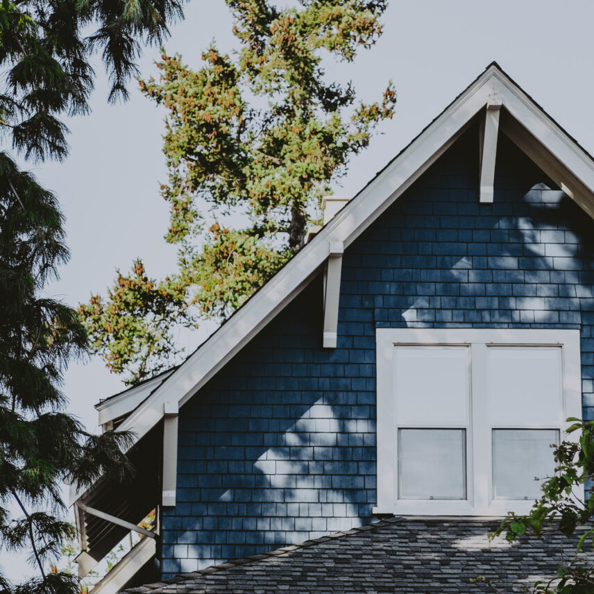 Close-up of a blue house exterior featuring white-trimmed windows and gabled roof surrounded by lush greenery