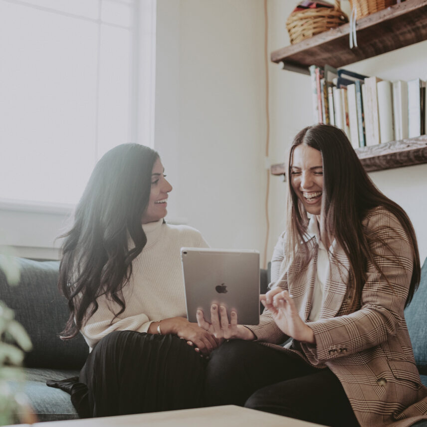 Colleagues sharing a moment of laughter while reviewing details on a tablet in a cozy living room setting, emphasizing a collaborative and approachable real estate experience