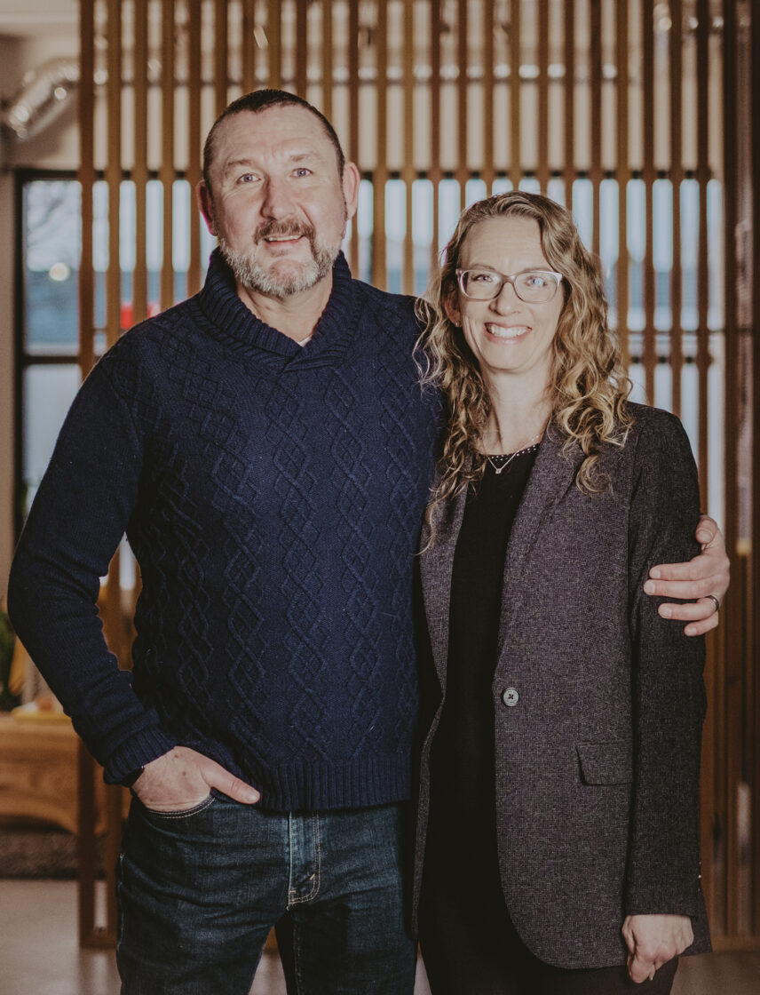 A man in a navy blue sweater and a woman in a dark blazer smiling together indoors