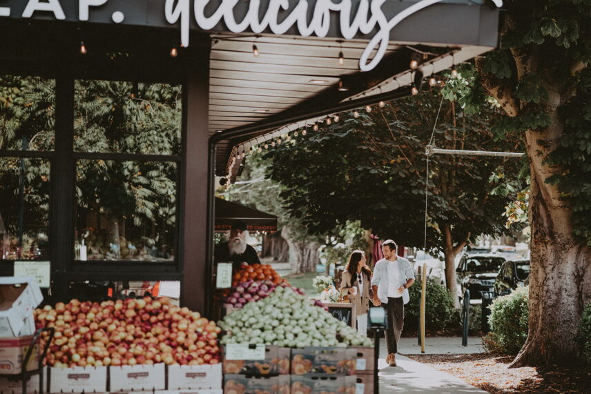 Street view in the Fairfield neighborhood near 624 Cornwall Street, Victoria, BC, featuring a local produce market with fresh fruit displays and people walking along the tree-lined sidewalk, managed by Folio Group at Clover Residential in Victoria, BC