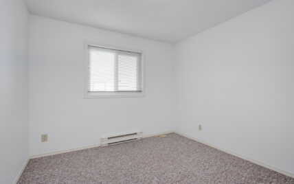 This image displays a clean, minimalistic bedroom at 1293 Layritz Place, managed by Clover Residential in Victoria, BC. The room features neutral carpeting, white walls, a window with blinds, and a baseboard heater, offering a blank canvas for personalization.