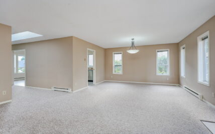 Open-concept living and dining area with large windows, carpeted flooring, and a central light fixture at 1293 Layritz Place, managed by Clover Residential in Victoria, BC