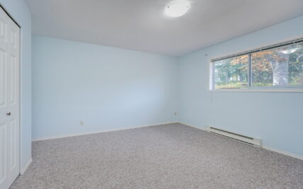 Spacious bedroom with light blue walls, large window overlooking greenery, and carpeted flooring at 1293 Layritz Place, managed by Clover Residential in Victoria, BC