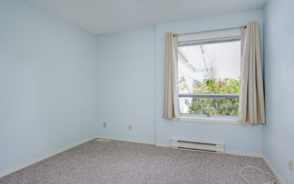 A cozy bedroom with soft blue walls, a window allowing natural light, and beige curtains at 1293 Layritz Place, managed by Clover Residential in Victoria, BC.