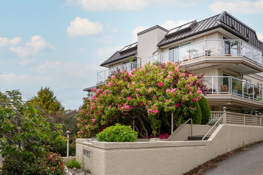 Exterior of a top-floor corner condo building in Fairfield, Victoria BC, with glass-railed decks and a blooming pink rhododendron
